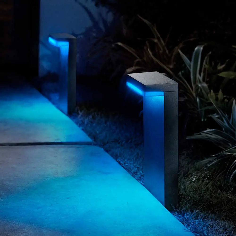 Baliza Nyro Philips Hue negro instalada en jardín iluminando camino exterior con luz azul y rosa.