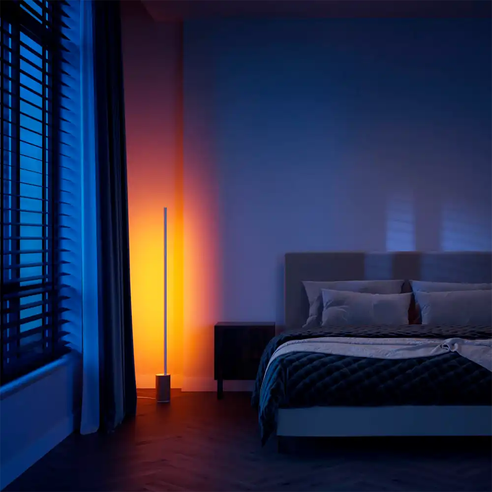 Lámpara de suelo Gradient Signe Philips Hue blanca y madera proyectando luz azul y naranja en dormitorio moderno